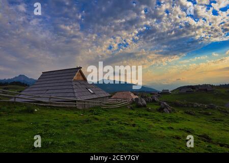 Holzhütte in Velika planina bei Sonnenaufgang. Schönes Foto der Agrarlandschaft mit Blick auf grünes Gras, Holzhaus und bewölktem Himmel. Stockfoto