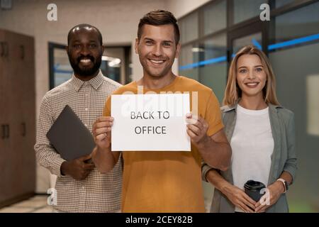 Junger glücklicher Kaukasier, Büroangestellter zeigt Papier mit Text ZURÜCK INS BÜRO vor der Kamera und lächelt, während er mit Kollegen im Büro steht Stockfoto