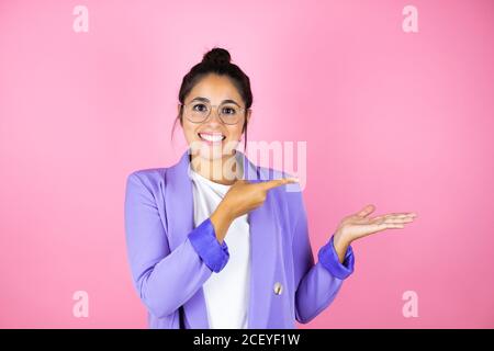 Junge schöne Geschäftsfrau über isolierten rosa Hintergrund lächelnd, zeigt und zeigt etwas, das auf ihrer Hand ist Stockfoto
