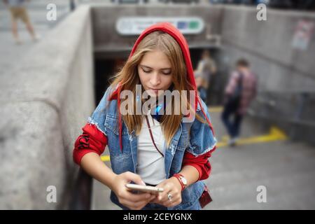 Frau im roten Hoodie liest Nachrichten auf dem Smartphone-Bildschirm, der in der Nähe der U-Bahn steht. Junge kaukasische Mädchen in der Nähe der U-Bahn-Station. Stockfoto