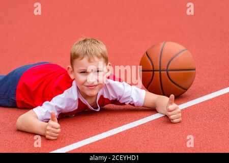 Junge posiert mit Basketball-Ball auf Kunstrasen rot. Sporttraining auf dem Feld. Kid Sport Online-Konzept Stockfoto