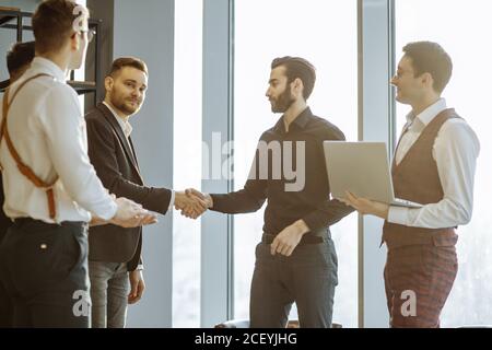 Zwei selbstbewusste Geschäftspartner, die nach einem erfolgreichen Meeting die Hände schütteln, nützliches Zusammenspiel zweier verschiedener Geschäftsteams, die in der Vorstandsetage versammelt sind. Erfolg Stockfoto