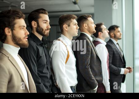 Seitenansicht auf junge kaukasische bärtige Geschäftsleute, die Seite an Fenster schauen, jeder in stilvollem elegantem Smoking. Isoliert in modernem Büro Stockfoto