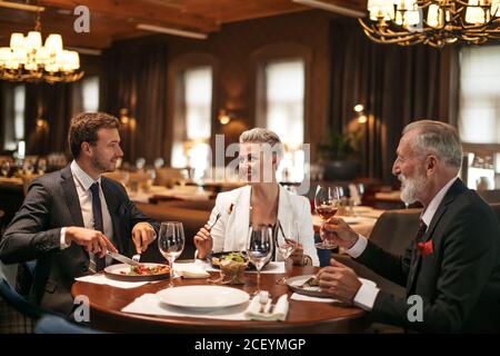 Gute fröhliche Freunde sitzen am runden Holztisch in gemütlichen weichen Stühlen im hell beleuchteten Restaurant, trinken Alkohol, essen leckeres Essen, lächeln glücklich, posieren o Stockfoto