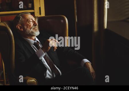 Gutmütiger alter Mann drückt Ruhe und positive Emotionen aus, sitzt bequem im dunklen Raum und schaut nachdenklich, horizontal abgeschossen Stockfoto