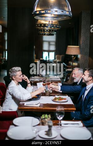 Zufriedene fröhliche Geschäftspartner in formeller Kleidung, sitzen am gedeckten Tisch, heben Weingläser mit fröhlichen Gesichtern auf, feiern leidenschaftlich den Geburtstag Stockfoto