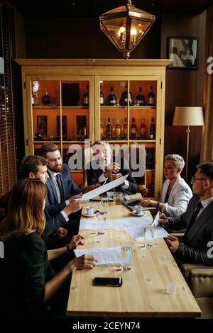 Team von jungen Fachleuten diskutiert Anzahl der wichtigen Fragen mit Ein älterer bärtiger Mann an einem langen Tisch im Büro mit Ledersesseln und Sty Stockfoto