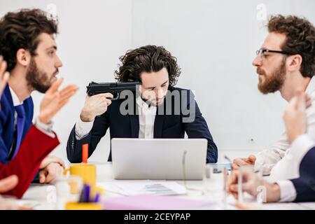 Junge kaukasische Mann in formeller Kleidung wollen Selbstmord begehen, während im Meeting-Büro mit fluchenden Kollegen sitzen, Probleme und Frist zu wor Stockfoto