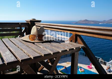 Strohhut mit Sonnenbrillen auf Holzterrasse des Urlaubs villa oder Hotel mit Stuhl Tisch mit Meer und Wimmen Blick auf den Pool Stockfoto