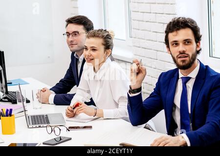 Positive Gruppe von Geschäftsleuten versammelten sich, um Geschäftsideen in einem modernen Sitzungssaal zu diskutieren, begeisterte und aktive Mitarbeiter sitzen zusammen auf dem Tisch Stockfoto