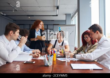 Sympathischer Geschäftskollegen in formeller Kleidung kamen zusammen, um das Geschäftsprojekt auf dem Tisch zu besprechen. Flipchart hinten, Laptops, Bleistifte auf dem Tisch. Stockfoto