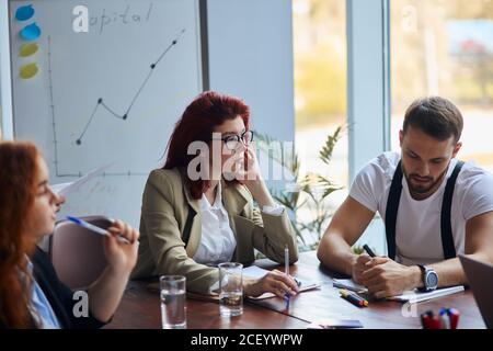 Frau mit roten Haaren Teamleiter sitzen Kollegen am Tisch mit jungen Geschäftskollegen zuhören, während sie sprechen. Seitenansicht auf Geschäftsfrau. Büro Stockfoto