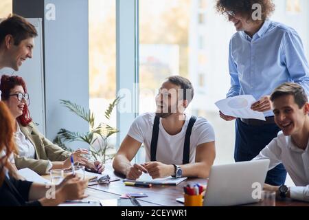 Team von fröhlichen kaukasischen Geschäftsleuten in eleganten formellen Kleidung sitzen zusammen am Tisch und diskutieren etwas, während lachen und lächeln. Alles gute für das Team Stockfoto