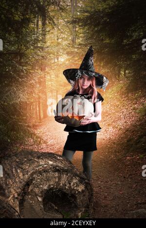 Eine junge Hexe hält einen Jack O Laterne Kürbis in einem magischen Wald in der Nacht. Halloween-Konzept. Stockfoto