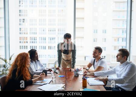 Selbstbewusster Mann, der junge freundliche, glückliche Führungskräfte unterrichtet, Coach, der mit Kollegen im modernen Büro mit Panoramafenster spricht Stockfoto