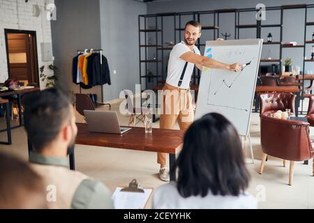 Selbstbewusster, enthusiastischer junger Coach hält eine Rede vor Kollegen, die ihm im Büro aufmerksam zuhören und legere Kleidung tragen Stockfoto