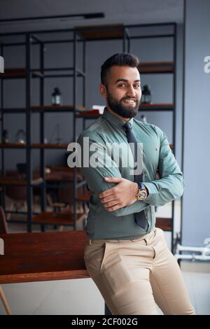 Bärtiger junger gutaussehender Geschäftsmann aus dem Nahen Osten, der im Büro steht, die Kamera anschaut, erfolgreicher Teamleiter, der formelle Kleidung trägt und für Fotos posiert Stockfoto
