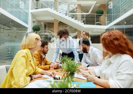Junge lustige Teamleiterin in lässiger Kleidung, hat Afro-Frisur, trägt runde Brille, lehnt sich vor positiven, fröhlichen Menschen auf den Tisch. Designer Stockfoto