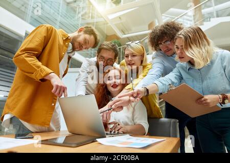 Unterer Schuss der fröhlichen Menschen mit dem charmanten Füchshaarigen Weibchen in der Mitte, stehen im modernen Büro, konzentriert im Laptop-Estrich, drücken freudige Expres aus Stockfoto