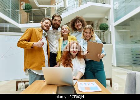 Gruppe von jungen kreativen Designern in legerer Kleidung, versammelt, um Fotos in hell beleuchteten modernen Büro zu machen, starren voraus mit breiten Lächeln, aus geschossen Stockfoto