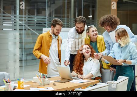 Ziemlich langhaarige Mädchen in weißer Bluse gekleidet, von fröhlichen Kollegen umgeben, Ausdruck freudigen Ausdruck, zeigt hervorragende Ergebnis der Arbeit Startup Stockfoto