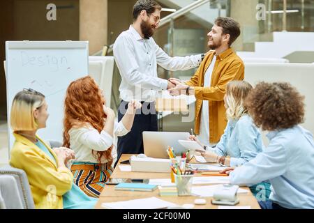 Edler tapferer Manager mit dickem Bart, in formalem Tuch schüttelt sich die Hände mit talentierter junger Designerin, legt Hand auf Schulter, gratuliert mit neuem Job-Position Stockfoto