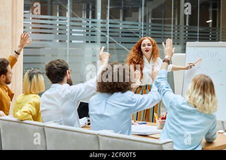 Charmante hübsche Trainerin steht vor jungen Designern, öffnet den Mund, überrascht von realem Interesse der Menschen, sieht erhobene Hände, rechte Seite Stockfoto
