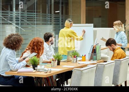 Team von jungen cleveren Designern im stilvollen Büro, blonde Erwachsene weibliche steht mit zurück in Richtung Kamera, schreibt wichtige Informationen über weiße Schwarzschweine Stockfoto