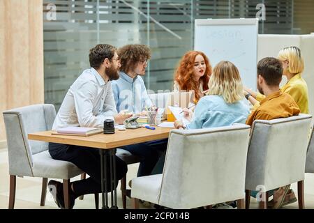 Fröhliche junge Kollegen, lässig gekleidet, diskutieren neues Design-Projekt, sitzen am Holztisch in modischen grauen Sesseln, beschäftigt bei der Arbeit, linke Seite Stockfoto