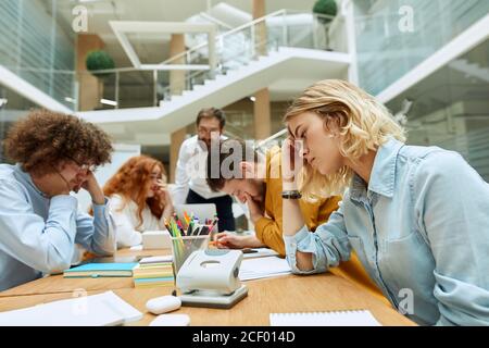 Verträumte junge Frau im hellblauen Hemd, Kopf halten, Stirn mit Fingern berühren, zur Seite schauen, über arbeitsbezogene Themen im modernen Coworking-Off nachdenken Stockfoto