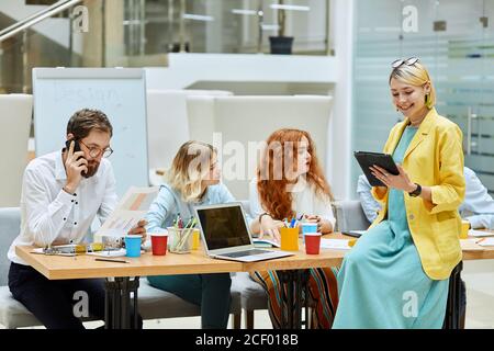 Team von fleißigen Designern arbeiten hart im modernen Büro, ziemlich charmante Frau sitzt auf dem Tisch mit Laptop an den Händen, Texte mit Freunden im Chat, sieht s Stockfoto