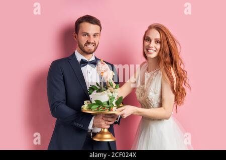 Lustige junge Paar hält süßen Kuchen isoliert über rosa Hintergrund. Mann im Smoking und Frau im weißen Hochzeitskleid Stockfoto