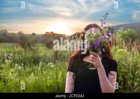 Junges Mädchen zu Fuß mit lila Wald Blumen in den Händen und Versteckt ihr Gesicht in Blumenstrauß vor dem Hintergrund des Sonnenuntergangs Stockfoto