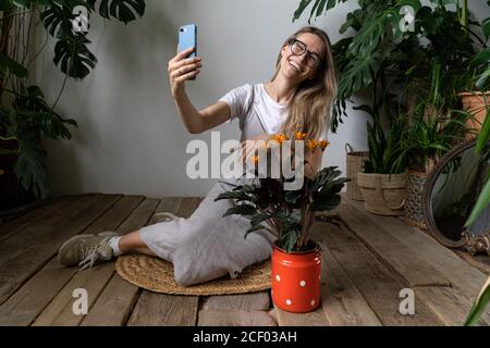 Glückliche Gärtnerin trägt Leinenkleid, lächeln und sprechen auf Video-Anruf auf dem Smartphone, sitzen auf Holzboden in der Nähe blühenden calathea Pflanze in o Stockfoto