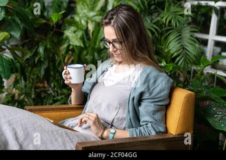 Die Gärtnerin trägt Leinenkleid, sitzt auf einem Stuhl im grünen Haus, ruht sich aus, benutzt Smartphone und trinkt Tee aus einer Tasse in Harmonie mit den Pflanzen ar Stockfoto