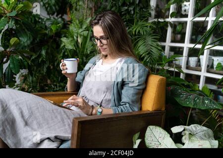 Die Gärtnerin trägt Leinenkleid, sitzt auf einem Stuhl im grünen Haus, ruht sich aus, benutzt Smartphone und trinkt Tee aus einer Tasse im Einklang mit den Pflanzen Stockfoto
