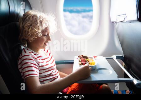 Kind auf dem Fenstersitz des Flugzeugs. Kinderflugmahlzeit. Kinder fliegen. Spezielles Bordmenü, Speisen und Getränke für Baby und Kind. Junge essen gesundes Mittagessen in der Luft Stockfoto