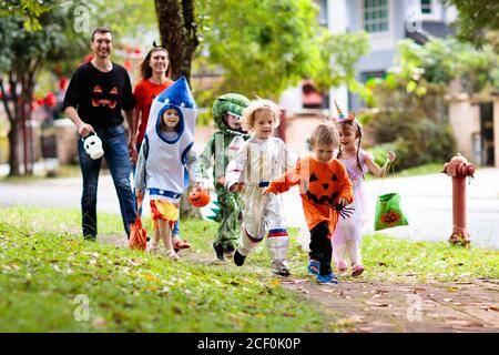 Kind in Halloween-Kostüm. Asiatische und kaukasische Kinder und Eltern mit gemischten Rassen tricksen oder gönnen sich auf der Straße. Kleiner Junge und Mädchen mit Kürbislaterne und Dose Stockfoto