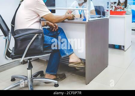 Mann und Frau Bankbeamte tragen Gesichtsmasken und haben Ein transparenter Schutzschirm zwischen den Transaktionen, um das zu verhindern Verbreitung des Corona Virus(C Stockfoto