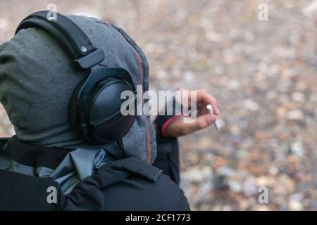 Stilvoller junger Kerl oder Hipster, der Musik in Kopfhörern mit Zigarette in der Hand hört. Blick von hinten. Entspannung im Freien. Stockfoto
