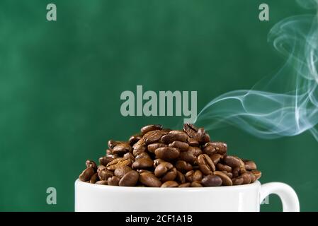 Internationaler Tag des Kaffees Konzept. Nahaufnahme weiße Kaffeetasse voller Kaffeebohnen auf grünem Hintergrund mit Rauch auf der Oberseite. Stockfoto