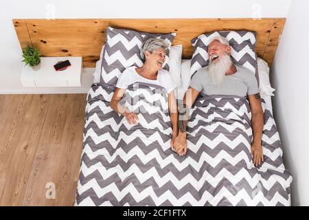 Glückliches Seniorenpaar, das Spaß hat und die Hände im Bett hält. Ältere Menschen lachen im Bett - Fokus auf Gesichter - Ältere fröhliche Lifestyle-Konzept Stockfoto