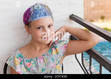 Lächelndes kaukasisches kleines Mädchen in bunten Kleid und Bandanna sitzt auf einem Balkon am Sommertag, Nahaufnahme im Freien Porträt Stockfoto