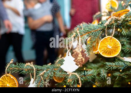 Getrocknete Orangenscheiben und baumförmige Lebkuchen, die an einem Weihnachtsbaum hängen, mit Weihnachtslichtern und Menschen im Hintergrund. Stockfoto