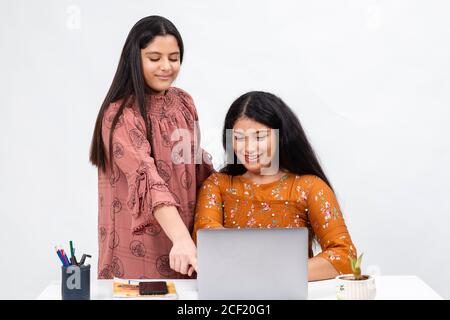 Zwei junge indische Mädchen suchen in einem Laptop. Ein Teenager sitzt auf einem Stuhl, während ein anderer über ihre Schulter in ihren Computer schaut. Stockfoto