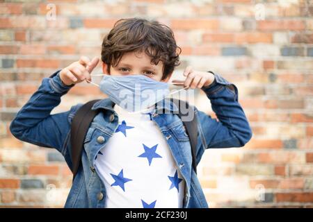 Kaukasisches Kind mit Schultasche auf eine Gesichtsmaske setzen. Er ist auf der Straße und geht nach der Quarantäne auf Covid-19 zur Schule. Stockfoto