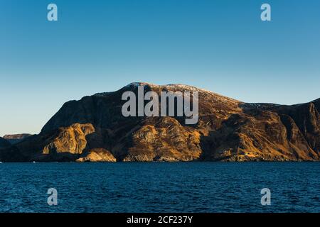 Steile Klippe an der Küste mit kleinen Häusern und Schnee Auf der Spitze in Norwegen an hellen sonnigen Tag von gesehen Boot Stockfoto