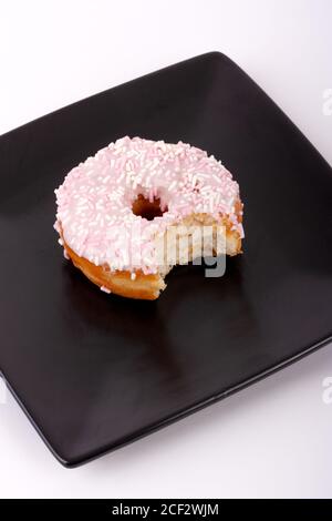 Milchdonut mit einem Biss aus ihm genommen, auf einem schwarzen quadratischen Teller, mit einem weißen Hintergrund. Stockfoto