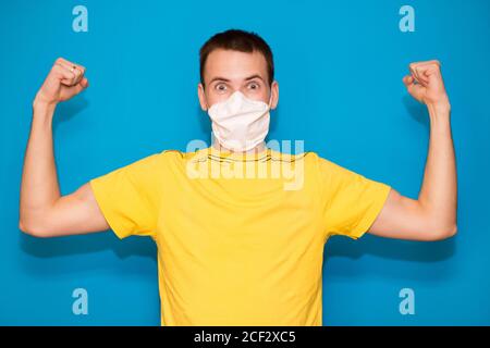 Junger gutaussehender Mann in medizinischer Maske und gelbem T-Shirt isoliert auf blauem Hintergrund. Starke Person zeigt Arm Muskel, zuversichtlich und stolz auf die Macht zu d Stockfoto