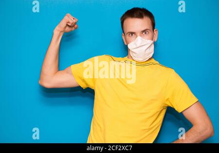 Junger gutaussehender Mann in medizinischer Maske und gelbem T-Shirt isoliert auf blauem Hintergrund. Starke Person zeigt Arm Muskel, zuversichtlich und stolz auf die Macht zu d Stockfoto